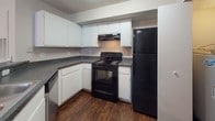 This single-floor unit is finished with dark hardwood-style flooring that runs consistently throughout the main living areas, bedrooms, and hallways, creating a cohesive and polished aesthetic. Textured ceilings are present throughout, complementing the neutral, light-toned walls.

The living area is generously proportioned, benefiting from natural light through vertical-blind-covered windows and a ceiling fan overhead. A partial-wall divider separates the living space from the kitchen, which features white cabinetry with dark countertops in an L-shaped configuration. Black appliances, including a refrigerator, electric range with overhead vent, and dishwasher, provide a clean contrast to the white cabinetry. A pantry closet is conveniently located adjacent to the kitchen.

The bedroom offers ample space with access to a dedicated walk-in closet, along with an additional door providing further storage or access. The bathroom includes a full tub-shower combination with white tile surround, a toilet, and built-in shelving, offering functional utility in a clean, straightforward layout. Hallway access connects the living and private areas of the unit seamlessly.