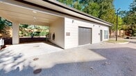 This purpose-built car wash facility is situated within the Tapestry Long Farm residential complex. The structure features a spacious covered area with a high ceiling, providing ample room for vehicle cleaning operations. The concrete floor is equipped with a drainage system, evident from the visible grates, ensuring proper water management. Adjacent to the covered area is a garage with a roll-up door, likely used for storage of cleaning supplies and equipment. The exterior is painted in a clean, neutral color scheme, harmonizing with the surrounding architecture. The facility is nestled in a landscaped environment, with mature trees and mulched areas providing a pleasant, natural backdrop. Red-colored car wash equipment is visible, indicating the presence of specialized cleaning apparatus. The location appears to be easily accessible, with paved driveways leading to and from the washing area. This well-designed amenity offers residents a convenient on-site solution for maintaining their vehicles in a serene, residential setting.