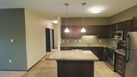 This single-floor space centers around a thoughtfully designed kitchen that serves as the focal point of the open layout. The kitchen is outfitted with rich, dark espresso-toned cabinetry that extends to the upper and lower walls, providing ample storage throughout. Granite countertops in warm, neutral tones run along the perimeter and are echoed on the substantial central island, which offers additional prep space and casual seating. Two elegant pendant lights hang above the island, complementing the flush-mount ceiling fixture nearby to create layered, warm illumination throughout the space. A stainless steel sink is positioned along the back counter, and black appliances, including a dishwasher, integrate seamlessly with the cabinetry. The flooring features neutral stone-look tile, adding texture and durability underfoot. Warm beige walls contrast tastefully with the darker cabinetry accents along the kitchen wall. A hallway visible from the kitchen leads to additional rooms, with dark-framed doorways maintaining a consistent design aesthetic throughout. The overall ambiance is warm, contemporary, and functional, with cohesive finishes that lend the space a polished, move-in-ready appeal.