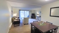 This single-floor space offers a well-organized, open-concept layout that seamlessly integrates the living, dining, and workspace areas into one cohesive environment. The living area is comfortably appointed with soft carpeting underfoot and features a large sliding glass door that spans much of the exterior wall, flooding the room with natural light and offering views of the surrounding outdoor scenery. A wall-mounted air conditioning unit ensures year-round comfort. Adjacent to the living area, a dedicated media and workspace zone includes a television stand and a compact desk setup, providing both entertainment and functional utility. The dining area, situated toward the front of the space, features a dark wood dining table with matching chairs, accented by a green table runner, creating a defined yet open eating area. The neutral, warm color palette throughout — from the cream-toned walls to the beige carpeting — contributes to a calm and inviting ambiance. Framed artwork on the walls adds a subtle decorative touch. The overall layout is practical and efficient, making excellent use of the available floor space while maintaining an airy, open feel throughout.