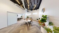 Located at 1300 S Casino Center Blvd in the heart of Las Vegas's vibrant Arts District, this modern office space offers a thoughtfully designed single-floor layout that balances open collaboration with private meeting functionality. The space is divided into two distinct areas: an open workstation zone and a dedicated conference room, seamlessly connected yet separated by a frosted glass sliding partition.

The open workspace features sleek white benching desks with privacy dividers, ergonomic rolling chairs, and abundant greenery that adds warmth to the clean aesthetic. Herringbone-patterned carpet tiles run throughout, lending a sophisticated yet approachable feel underfoot. Exposed black ceilings with visible ductwork and linear track lighting create an industrial-modern contrast against the crisp white walls, which are accented by a painted white brick lower half.

The conference room accommodates a full-sized meeting table surrounded by grey and gold upholstered chairs, with a wall-mounted display screen for presentations. A globe chandelier adds an elegant focal point overhead. High, horizontally positioned clerestory windows in both areas allow natural light to filter in while maintaining privacy. The overall ambiance is polished, professional, and purposefully curated for a productive work environment.