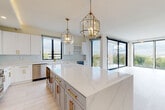 This elegantly appointed single-floor residence showcases a cohesive contemporary design with high-end finishes and a bright, open layout. The heart of the home is a stunning open-concept kitchen and living area, where large-format light tile flooring flows seamlessly throughout. The gourmet kitchen features a substantial center island topped with striking white marble-veined quartz, complemented by a two-tone cabinet scheme of crisp white uppers and soft blue-gray lower island cabinetry, all adorned with brushed gold hardware. A professional-grade stainless steel range, built-in dishwasher, farmhouse sink, wine refrigerator, and a glass-tile subway backsplash round out the chef's kitchen. Geometric brass pendant lights add warmth and visual interest overhead. The expansive living area is defined by floor-to-ceiling black-framed sliding glass doors that invite abundant natural light and offer sweeping panoramic views of the surrounding treetop and cityscape. The primary bathroom features a double-sink white vanity, dual round gold-framed mirrors, gold fixtures, and a glass-enclosed walk-in shower with marble tile surround. A well-organized walk-in closet with built-in shelving, hanging rods, and gold-handled drawers provides generous storage. Additional bedrooms are bright and well-proportioned, maintaining the home's clean, modern aesthetic.
