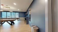 This well-appointed recreational space combines entertainment and relaxation into a single, cohesive environment. The room is finished with striking deep blue walls that extend throughout, complemented by white wainscoting panels along the lower portion of the walls, creating a polished, two-tone aesthetic. Light wood-look flooring runs throughout, adding warmth and continuity to the space.

The layout is divided into distinct functional zones. A game area features a full-size pool table with overhead pendant lighting and a wall-mounted cue rack, alongside billiards-themed wall art. Adjacent to it, a bar counter with bar stools provides a casual gathering spot. Large floor-to-ceiling windows with blackout curtains line one wall, allowing natural light and outdoor views while maintaining privacy when needed.

The lounge area is furnished with a plush blue sofa, rounded cognac leather armchairs, and a round marble-top coffee table set atop a geometric patterned rug. Three wall-mounted flat-screen televisions are arranged side by side on one wall, making this section ideal for viewing. Recessed lighting throughout ensures consistent, well-distributed illumination. The overall ambiance is modern, inviting, and designed for social interaction.