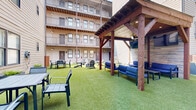 Located in the heart of Nashville's 12 South neighborhood, this shared outdoor courtyard at 1016 18th Ave S offers a well-designed communal amenity space nestled within a modern multi-story residential building. The courtyard is fully enclosed by the surrounding structure, creating a private and sheltered atmosphere while remaining open to natural light throughout the day.

The ground is finished with lush artificial turf, providing a clean, low-maintenance surface ideal for outdoor gatherings and casual recreation. A central fire pit with Adirondack-style seating serves as a natural gathering point, offering a relaxed social hub for residents. Additional outdoor dining furniture, including a table-and-chair set, is positioned along the perimeter, accommodating both small group meals and leisure activities.

Raised planter boxes add greenery and visual interest throughout the space, complementing the turf with natural elements. A warm-toned horizontal wood privacy partition provides a defined boundary within the courtyard, adding architectural texture. A covered pergola structure toward one end offers a shaded retreat. The exposed beam ceilings of the building's covered walkways and balconies overhead contribute an industrial-modern aesthetic, with metal railings lining each floor. The courtyard's design balances functionality and comfort, making it a versatile outdoor living extension for the community.