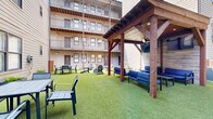 Located in the heart of Nashville's 12 South neighborhood, this shared outdoor courtyard at 1016 18th Ave S offers a well-designed communal amenity space nestled within a modern multi-story residential building. The courtyard is fully enclosed by the surrounding structure, creating a private and sheltered atmosphere while remaining open to natural light throughout the day.

The ground is finished with lush artificial turf, providing a clean, low-maintenance surface ideal for outdoor gatherings and casual recreation. A central fire pit with Adirondack-style seating serves as a natural gathering point, offering a relaxed social hub for residents. Additional outdoor dining furniture, including a table-and-chair set, is positioned along the perimeter, accommodating both small group meals and leisure activities.

Raised planter boxes add greenery and visual interest throughout the space, complementing the turf with natural elements. A warm-toned horizontal wood privacy partition provides a defined boundary within the courtyard, adding architectural texture. A covered pergola structure toward one end offers a shaded retreat. The exposed beam ceilings of the building's covered walkways and balconies overhead contribute an industrial-modern aesthetic, with metal railings lining each floor. The courtyard's design balances functionality and comfort, making it a versatile outdoor living extension for the community.
