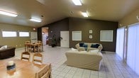 This single-story space is defined by its vaulted ceilings, which lend an airy, open quality to the interior. The main living area is generously proportioned, featuring light tile flooring throughout that contributes to a clean, cohesive aesthetic. Deep brown-toned walls provide a warm, grounding contrast against the lighter furnishings and flooring. Overhead, flush-mounted fluorescent lighting ensures the space remains well-lit throughout.

The living area is anchored by a pair of cream-colored sofas accented with teal and navy throw pillows, positioned to encourage conversation. Framed art pieces are mounted on the walls, adding a modest decorative touch. Windows with blinds allow for natural light control while maintaining privacy.

Adjacent to the living area, a dining space accommodates a natural wood table and matching chairs, offering a casual and functional setting for meals. A doorway leads into a kitchen area, where a white refrigerator and cabinetry are visible in the background. A tall potted plant near the transition between spaces adds a natural element. The overall layout is open and practical, designed to maximize usability across the living, dining, and kitchen zones.