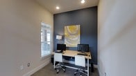 This single-story space is designed as a functional and stylish workspace. The room features a striking dark slate-blue accent wall that serves as a bold focal point, contrasting effectively against the light neutral tones of the surrounding walls. A vibrant abstract painting in yellow, white, and grey hangs prominently on the accent wall, adding an artistic touch that enlivens the environment.

The workspace is equipped with a shared light wood desk that accommodates two workstations, each outfitted with a desktop monitor, keyboard, and mouse. Two modern white rolling office chairs provide comfortable seating. A sleek desk lamp offers supplemental task lighting alongside recessed ceiling lights that ensure the room is well-illuminated.

A window fitted with horizontal blinds allows natural light to filter into the space, contributing to a bright and inviting atmosphere while maintaining privacy. The floor is covered with neutral-toned carpet, adding warmth and sound absorption to the room. Clean white baseboards and trim complete the polished aesthetic. Overall, the space balances practicality with a contemporary design sensibility, making it an efficient and visually appealing work environment.