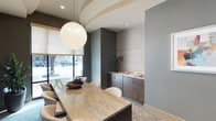 This elegantly designed single-floor space exudes a refined, contemporary aesthetic that balances form and function. At the heart of the space sits a substantial marble-topped table with a rich, veined surface, surrounded by sleek white chairs that invite gathering and conversation. A large, luminous globe pendant light serves as a dramatic focal point, casting a warm glow over the room and complementing the layered ceiling detail above.

The design palette is thoughtfully curated, combining cool blue-grey walls with warm wood cabinetry and a textured grasscloth-style accent wall that adds depth and visual interest. A built-in sideboard with dark wood cabinetry provides both practical storage and a stylish display surface, topped with decorative accents including a tall vase with greenery and a small bowl.

Floor-to-ceiling windows along one wall flood the space with natural light and offer views of the surrounding streetscape, connecting the interior to the outdoors. Underfoot, a patterned area rug with a flowing botanical motif adds a soft, artistic touch to the polished overall composition. Recessed lighting throughout ensures the space is well-illuminated at all times. The result is a polished, welcoming environment ideal for meetings or social gatherings.