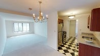 This single-floor residence is thoughtfully laid out to balance open living areas with functional private spaces. The living and dining areas are connected via a subtle split-level design, unified by plush wall-to-wall carpeting and bright white walls that promote a clean, airy atmosphere. A gold candelabra-style chandelier serves as a focal point in the dining area, adding a classic decorative touch. A large window with vertical blinds allows natural light to filter into the living space, offering views of the surrounding greenery.

The kitchen makes a bold statement with its striking black-and-white checkered tile flooring. Rich wood cabinetry with brass hardware provides ample storage, complemented by granite-style countertops and a full suite of stainless steel appliances, including a gas range, built-in microwave, and refrigerator. A walk-in closet is visible from the kitchen corridor, adding practical storage.

The bedrooms are carpeted and generously proportioned, each featuring large windows with vertical blinds that bring in natural light. One bedroom has direct access to a bathroom with granite countertops and a tub-shower combination. A second bathroom features cheerful checkered tile flooring and a tub-shower with decorative curtains.