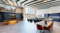 This generously sized two-floor amenity space is defined by its dramatic double-height main hall, where exposed beam ceilings painted in deep green contrast against bright white walls. Large-format porcelain tile floors and floor-to-ceiling sliding glass doors flood the ground level with natural light and provide direct views of an outdoor courtyard. The open layout accommodates distinct functional zones: a plush lounge area furnished with teal velvet seating, a full-size pool table positioned beneath the grand staircase, and a long communal dining table anchored near a fully equipped demonstration kitchen. The kitchen features bold navy cabinetry with brass hardware, a shimmering mosaic tile backsplash, and stainless steel appliances. A built-in navy shelving unit near the entrance provides curated display and storage. A dedicated media nook houses a large-screen display flanked by wall sconces and complementary seating. The sculptural staircase, adorned with a bold geometric mural, connects to the upper level, which hosts a co-working suite. This workspace includes a large conference table, private glass-enclosed collaboration pods, and a vibrant geometric carpet, delivering a dynamic and functional environment throughout.