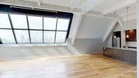 This distinctive top-floor loft showcases exceptional architectural character through its dramatic angled skylight wall, which floods the open living area with abundant natural light and frames sweeping views of the surrounding cityscape and treetops. Exposed white-painted structural beams traverse the ceiling, lending an industrial-chic aesthetic that complements the warm light hardwood flooring throughout the main living spaces.

The open-plan living and kitchen area is anchored by a well-appointed kitchen featuring rich dark wood cabinetry, a classic white subway tile backsplash, stainless steel appliances, and a generous granite-topped island with an integrated sink and pendant lighting overhead. Large multi-pane windows further extend the connection to the urban panorama beyond.

The bedroom continues the angled glazing motif, offering its own treetop outlook and plush carpeted flooring. A dedicated walk-in closet provides ample storage with built-in shelving and hanging rails. An additional carpeted bedroom with its own walk-in closet is also present, serviced by exposed ductwork at the ceiling.

The bathroom is finished with large-format tile flooring, a granite vanity top, and dark wood cabinetry consistent with the kitchen's palette. A dedicated in-unit laundry room houses a full-size side-by-side washer and dryer, completing this thoughtfully designed urban loft.