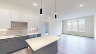 Located at 8680 State Line Rd in Leawood, KS, this thoughtfully designed one-bedroom, two-bathroom residence offers a clean, contemporary aesthetic throughout. The open-concept kitchen and living area serves as the heart of the home, featuring sleek two-tone cabinetry with white upper cabinets and slate-gray lower cabinets, a neutral tile backsplash, and a full suite of stainless steel appliances. A center island with a white quartz countertop provides additional prep space and casual seating, while stylish cylindrical pendant lights add a modern touch overhead. Light-toned hardwood-style flooring flows through the main living area, which is bathed in natural light from large windows and a glass door opening to a private balcony with outdoor views. The generously sized primary bedroom features plush carpeting, a ceiling fan, and direct balcony access via sliding glass doors. It is complemented by a spacious walk-in closet outfitted with wire shelving on all sides, offering excellent storage capacity. Two bathrooms serve the residence, finished with light wood-look flooring and clean white fixtures. The overall palette of crisp whites and warm neutrals creates a bright, airy ambiance throughout this well-appointed home.