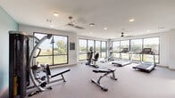 This single-level fitness center is thoughtfully divided into two interconnected zones, separated by a set of sleek sliding barn-style doors with frosted glass panels. The main cardio and strength area features large floor-to-ceiling windows along two walls, flooding the space with natural light and offering open views of the surrounding landscape. Ceiling fans and recessed lighting ensure consistent comfort throughout. Equipment in this zone includes treadmills, an elliptical, a recumbent bike, a multi-function cable machine, adjustable benches, and a full dumbbell rack, catering to a wide range of training needs. Durable, low-pile carpet covers the floors throughout.

The secondary zone, accessed through the barn doors, functions as a dedicated studio and functional training area. It features a full-length mirrored wall paired with a large mounted smart TV, ideal for guided workouts. This space also houses stability balls, foam rollers, step platforms, and additional accessories for group or solo training. A bold teal accent wall adds visual character to a corner strength station equipped with a multi-function weight machine. Large windows in this section continue the theme of natural light and outdoor connectivity, creating an energizing environment throughout the entire facility.