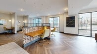 Located at 500 W 14th St in Richmond, Virginia, this thoughtfully designed single-floor community space blends contemporary style with functional versatility. The open-plan layout is anchored by herringbone-patterned hardwood flooring that flows throughout, creating visual continuity across each distinct zone.

The coworking area serves as the heart of the space, featuring a standout workstation built with exposed brick bases and warm wood surfaces, complemented by gold-accented rolling chairs. Mesh partitions provide subtle separation without sacrificing the open feel. Large black-framed windows line the perimeter, flooding the interior with natural light and offering views of the surrounding Richmond streetscape.

A dedicated lounge area offers a relaxed retreat, furnished with tan leather armchairs, a soft grey sofa, and a statement drum pendant light in brushed gold. A wall-mounted flat-screen television adds to the comfort of this gathering spot.

A private dining or conference room rounds out the space, enclosed by glass walls and featuring floor-to-ceiling corner windows. A rustic wood table surrounded by plush blue velvet chairs sits beneath a striking sputnik chandelier, making it ideal for meetings or intimate gatherings. Contemporary artwork accents the walls throughout, and recessed lighting ensures a well-lit environment at all hours.