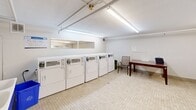 Located at 280 Dundas Street East in Toronto, this shared laundry facility offers a clean, functional space designed to meet the everyday needs of building residents. The room is well-lit by overhead fluorescent lighting that illuminates the bright white walls and classic mosaic tile flooring throughout.

The main laundry area is lined with a row of four front-loading washing machines and four top-loading dryers, all equipped with card or coin-operated payment systems for easy use. A large wall mirror above the machines adds a sense of openness to the space. A small table and chair are positioned along one wall, providing a comfortable spot for residents to wait while their laundry cycles complete.

An adjacent utility area features an industrial-style deep sink workspace, ideal for hand-washing or pre-treating garments, along with recycling bins to encourage responsible waste management. This secondary section is accessed through a solid door and includes exposed overhead piping, giving it a practical, utilitarian character.

Ventilation infrastructure is visible along the ceiling, ensuring proper airflow throughout the facility. The overall layout is efficiently organized, making it easy to navigate and use. This well-maintained laundry room provides a convenient and accessible amenity for all residents of the building.