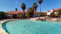 Located on Beechnut Street in Houston, Texas, this single-story residential community offers a well-rounded set of shared amenities designed for everyday comfort. The outdoor area is a standout feature, showcasing a large resort-style swimming pool with crystal-clear turquoise water, a natural rock waterfall formation, and mature palm trees that lend a tropical ambiance under the open Texas sky. The warm earth-toned exterior of the buildings, accented by pitched rooflines and traditional architectural detailing, frames the pool area and creates an inviting communal atmosphere.

The common entry corridor serves as the primary access hub for residents, featuring textured white paneled walls and durable tile flooring underfoot. A wall-mounted bank of individual metal mailboxes, framed in a bold red trim and lit by a classic lantern-style fixture, provides a centralized mail retrieval point for all residents. The corridor opens on both ends, allowing natural light to flow through and offering sightlines to the surrounding landscaped grounds.

Security is emphasized throughout the property, with a prominent black metal security gate controlling access between the entry corridor and the interior pathways, which are lined with brick pavers and lush tropical plantings. This community combines practical amenities with an outdoor-oriented lifestyle in a well-established Houston neighborhood.