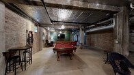 Located at 1508 Madison Ave in Memphis, TN, this distinctive entertainment space masterfully blends industrial architecture with eclectic, inviting design. The single open floor plan offers generous room to move and gather, anchored by original exposed brick walls that lend warmth and historic character throughout. Overhead, embossed tin ceiling tiles add an ornate contrast to the exposed black iron pipes, ductwork, and structural concrete columns that underscore the space's raw, industrial aesthetic. Polished concrete floors provide a durable foundation beneath the varied activity zones. A full-sized billiards table serves as a central focal point, complemented by a lounge area furnished with a tufted leather sofa, plush velvet armchairs, and a bold Persian-style area rug. Vintage-style posters and wall art punctuate the brick surfaces, adding cultural flair. A dedicated game nook features a large wall-mounted Scrabble board with casual bean bag seating, ideal for relaxed play. A TV wall with sconce lighting and a high-top bar table offer additional entertainment options. Small windows along the brick walls allow for modest natural light, while pendant and accent lighting create a warm, intimate ambiance throughout. This space is well-suited for social gatherings, private events, or recreational use.