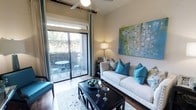 This thoughtfully designed single-floor space makes a strong impression through its cohesive interior styling and functional layout. The living area serves as the heart of the home, showcasing warm hardwood flooring that anchors the room with a sense of comfort and elegance. A large sliding glass door floods the space with natural light and provides direct access to a private balcony, where a bistro table and chairs create an inviting spot for outdoor relaxation. The balcony is framed by lush greenery, adding a refreshing natural backdrop. Inside, the living room is furnished with a classic white sofa adorned with bold teal and patterned throw pillows, complemented by a sleek dark wood coffee table as a striking centerpiece. A large abstract painting in greens, blues, and purples adds an artistic focal point to the wall, tying together the vibrant color palette. A ceiling fan with warm-toned lighting enhances airflow and ambiance throughout the space. Neutral wall tones provide a calm, versatile canvas that allows the teal accents and decorative elements to stand out. Overall, the space blends contemporary style with a welcoming warmth, making it both visually appealing and highly livable.