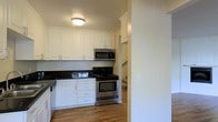 This two-story space offers a functional and clean aesthetic, highlighted by a well-designed kitchen visible from the entry. The kitchen features crisp white shaker-style cabinetry that extends to the upper walls, providing generous storage capacity. Dark granite or stone countertops contrast sharply with the white cabinets, creating a classic and visually appealing combination. Stainless steel appliances, including a gas range, over-the-range microwave, and dishwasher, lend a modern and practical character to the space. The warm hardwood-style flooring runs throughout, adding continuity and a welcoming feel underfoot. A staircase visible in the background connects the two floors, suggesting a thoughtful layout that separates living areas across levels. The ceiling features a flush-mount light fixture, maintaining a clean and uncluttered overhead profile. The textured ceiling adds subtle visual interest. Overall, the space presents a tidy, move-in-ready environment with a neutral palette that allows for versatile interior styling across both floors.