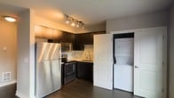 This well-appointed single-floor space showcases a thoughtfully designed kitchen that serves as the centerpiece of the unit. The kitchen features bold, dark cabinetry that contrasts beautifully against a light stone tile backsplash, creating a striking modern aesthetic. Stainless steel appliances, including a full-size refrigerator, electric range, built-in microwave, and dishwasher, offer a complete and functional cooking environment. A sleek sink with a modern gooseneck faucet is positioned beneath ample counter space, making meal preparation both practical and stylish. Track lighting mounted on the ceiling provides focused, adjustable illumination throughout the kitchen area, enhancing the contemporary feel. Dark hardwood-style flooring runs throughout, grounding the space with warmth and continuity. A standout convenience feature is the in-unit stacked washer and dryer, neatly housed behind a classic paneled door, keeping laundry accessible without compromising the overall aesthetic. The clean lines, cohesive color palette, and efficient use of space come together to create a modern, functional living environment suited for everyday comfort.