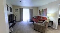 This single-floor residence offers a practical and well-organized layout across several interconnected rooms. The living room serves as the central gathering space, featuring plush carpeted flooring, a textured ceiling, and a large sliding glass door framed by floor-length grey curtains that allow generous natural light to fill the room. The space comfortably accommodates a seating arrangement with a sofa, accent chair, and coffee table, creating an inviting atmosphere. Wall-mounted artwork adds a decorative touch to the neutral-toned walls.

The floor plan includes two bedrooms, each furnished with beds and storage pieces, and benefiting from window access that brings in additional natural light. Hardwood-style flooring is visible in the central and transitional areas of the home, contrasting with the softer carpeted surfaces in the living and sleeping spaces. A dining or multipurpose area is situated near the kitchen zone, providing a practical space for everyday use.

Overall, the residence presents a cohesive and functional design with warm, neutral tones throughout, offering a comfortable and livable environment across its single level.