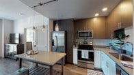 This contemporary single-floor residence combines modern design with functional living across several well-defined spaces. The kitchen is a standout feature, equipped with stainless steel appliances including a French door refrigerator, range, and over-the-range microwave. Two-tone cabinetry pairs dark upper cabinets with lighter lower units, complemented by a tile backsplash and a light-colored countertop island with pendant lighting overhead — ideal for both meal prep and casual dining.

The living area offers a relaxed, open atmosphere with access to a balcony, allowing natural light to fill the space. A dedicated office or den is separated from the main living area by a striking rustic barn door, providing privacy while maintaining an open feel. This room features large windows with urban views and ample workspace.

The bedroom is bright and serene, with wood-look flooring and a window that admits natural light. Two bathrooms serve the residence: one is a spacious, accessibility-friendly full bath with grab bars and generous floor space, while the second features a walk-in shower area, granite-topped vanity, and a layout that connects to additional storage. Wood-look flooring runs consistently throughout the unit, tying all spaces together with a warm, cohesive aesthetic.