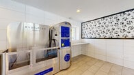 Located at 4673 Lake Valley Drive in Lisle, Illinois, this single-floor pet grooming facility is purpose-built to provide a clean, convenient, and welcoming environment for pet owners and their dogs. The space centers around a stainless steel Evolution dog wash station, a self-serve unit equipped with shampoo, rinse, blow dry, conditioner, and disinfectant functions, making it a fully featured grooming solution in a compact footprint.

The interior is finished with large-format light beige tiles laid in a herringbone-inspired pattern, contributing to a bright and hygienic aesthetic. White large-panel wall tiles line the perimeter, giving the room a modern, easy-to-clean surface throughout. A built-in countertop with a granite surface provides a practical workspace alongside the wash station, while a sturdy bench offers a convenient spot for pet owners to prepare their animals.

Natural light filters in through a window positioned above the counter area, keeping the space feeling open and airy. A playful paw print wall mural adds a whimsical, pet-friendly touch to the otherwise clean and minimal design. Upper glass panels and mirrors visually expand the space, and recessed lighting along with pendant fixtures ensure consistent illumination. A glass entry door offers views to the surrounding community, reinforcing the accessible, community-oriented nature of this well-appointed grooming amenity.
