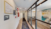 This single-floor commercial space presents a thoughtfully designed interior that balances modern aesthetics with functional practicality. The main corridor serves as the spine of the layout, finished with warm wood-look flooring and a dark runner rug accented with gold star motifs, guiding visitors through the space. Recessed lighting runs the length of the hallway, ensuring consistent, well-distributed illumination throughout.

One of the most distinctive architectural features is the floor-to-ceiling steel-framed glass partition wall lining one side of the corridor, offering visibility into an adjacent room furnished with a dining or conference table and an eye-catching orange pendant light. This transparency creates an open, connected atmosphere while maintaining defined zones. The opposite wall features white paneling with mounted utility and safety equipment, including fire alarm components, adding a functional but unobtrusive presence.

Exposed concrete columns punctuate the corridor, lending an industrial character that contrasts effectively with the clean white walls. A stainless steel elevator provides accessibility within the space. At the far end of the corridor, a glass-paneled entrance door draws in natural light from outside. A compact utility area with a built-in white countertop rounds out the practical offerings of this well-appointed commercial space.