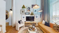 Located at 96 E Catawba St in Belmont, NC, this industrial-chic loft seamlessly blends historic character with contemporary design. The single-floor layout is anchored by dramatically tall ceilings with exposed wood beam details and visible ductwork, evoking a classic converted mill aesthetic. Original brick and reclaimed wood columns punctuate the open-concept living area, adding warmth and texture to the bright, airy space.

The living area is bathed in natural light from floor-to-ceiling windows dressed with teal curtains, and features warm hardwood flooring throughout. A curated gallery wall with an eclectic mix of framed artwork and decorative elements surrounds the media console, while a statement pendant light anchors the seating arrangement.

The kitchen is well-appointed with white shaker cabinetry, subway tile backsplash, granite countertops, stainless steel appliances including a full-size refrigerator, and open shelving for a modern, functional feel.

The bedroom offers a calm retreat with high ceilings, hardwood floors, warm-toned furnishings, and an arched full-length mirror adding architectural elegance. The bathroom features white square tile surrounding a full tub and shower, matte black fixtures, granite countertops, and gold hardware accents — a refined contrast of bold and soft finishes.