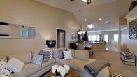 This single-floor residence showcases an open-concept layout that connects the living, dining, and kitchen areas into one cohesive, flowing space. The living room is anchored by a generously sized sectional sofa adorned with decorative pillows in neutral and grey tones, complemented by a wooden coffee table with fresh floral accents. A striking black chandelier-style lamp adds a touch of elegance alongside recessed lighting overhead.

The soaring vaulted ceilings are a defining architectural feature, creating an airy, expansive atmosphere throughout. A ceiling fan is centrally positioned to enhance comfort. Warm, neutral-toned walls and light carpeting contribute to the cohesive, understated palette.

Transitioning seamlessly from the living space, the dining area accommodates a full table and chairs beneath a classic chandelier. Beyond, the kitchen features dark cabinetry, a kitchen island, and large windows that allow natural light to fill the space. A hallway visible from the living room provides access to additional areas of the home. Overall, the space exudes a clean, welcoming ambiance ideal for everyday living and entertaining.