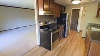 This single-floor space features a functional kitchen designed with a practical layout and quality finishes throughout. The kitchen is outfitted with rich, dark wood cabinetry providing ample overhead storage, complemented by a decorative mosaic tile backsplash that adds visual interest to the cooking area. A stainless steel electric range with a coil cooktop serves as the centerpiece, paired with a matching refrigerator and an under-cabinet range hood for ventilation. The flooring transitions from warm, light-toned laminate wood planks in the kitchen to plush carpeting in the adjacent living area, clearly delineating the two spaces while maintaining a cohesive feel. A ceiling-mounted light fixture provides overhead illumination, and a doorway visible in the background hints at additional rooms beyond. The overall ambiance is cozy and utilitarian, with the contrast between the dark cabinetry and lighter flooring creating a balanced aesthetic. The open sightline between the kitchen and carpeted area enhances the sense of connectivity within the single-level layout.