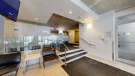 The lobby at 3400 Boul St-Elzéar O in Laval presents a contemporary, well-appointed entrance space designed to efficiently welcome and orient visitors and residents. The layout is organized across a split-level configuration, with the lower entry level connected to an elevated interior hallway by a short staircase and a glass-paneled railing system with polished stainless steel accents, lending the space a clean, modern aesthetic.

The lower entry level features large glass doors and expansive windows that flood the area with natural light and provide clear views of the surrounding streetscape. Light tile flooring and dark carpet entry mats define transition zones throughout. A seating area with sleek black leather chairs and a small side table is positioned near the windows, offering a functional waiting area.

Moving up the steps to the elevated section, the space opens to reveal two elevator banks, an exposed concrete structural column, and a textured feature wall that adds visual depth. The concrete ceiling and exposed beams contribute an industrial character, contrasted by recessed lighting and white painted walls. A bank of mailboxes and an intercom system are integrated along one wall. The corridor leading further into the building is carpeted in dark tones, maintaining a professional and practical finish throughout.