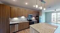 This contemporary single-floor residence is thoughtfully designed with an open-concept layout that seamlessly connects the kitchen and living areas. The kitchen serves as a central highlight, showcasing warm wood-toned cabinetry extending to the ceiling, granite countertops, a full suite of stainless steel appliances including a dishwasher, electric range, microwave, and refrigerator, and a spacious island with matching granite surface. Stylish pendant lighting hangs above the island, complementing the recessed ceiling lights throughout. Wood-look plank flooring flows consistently through the living spaces, adding warmth and visual continuity. The living area is bright and airy, featuring a ceiling fan and large sliding glass doors that open to a private balcony with exterior views. A secondary bedroom also benefits from generous window placement, allowing ample natural light. The bathroom is well-appointed with granite vanity countertops, wood-toned cabinetry, a tub/shower combination, and a large wall mirror. The bedroom area includes access to a dedicated closet and a full bathroom, providing practical organization and convenience. Neutral gray wall tones throughout the residence create a clean, cohesive aesthetic that complements the modern finishes.