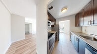 This single-floor residence offers a thoughtfully designed layout with distinct living zones connected by a central corridor. The galley-style kitchen is a functional highlight, featuring rich dark-walnut cabinetry lining both walls, light stone countertops, and a classic white subway tile backsplash. Stainless steel appliances — including a refrigerator, electric range, built-in microwave, and dishwasher — are integrated seamlessly, while large-format ceramic tile flooring completes the space. The kitchen opens directly into the living area, creating a natural flow throughout the unit.

The living area is bright and airy, benefiting from an expansive wall of floor-to-ceiling windows and a sliding glass door that opens to a balcony with open city views. Natural light streams in generously, highlighting the warm hardwood flooring that extends through the living and sleeping areas. Baseboard heating runs along the exterior walls, providing consistent comfort throughout.

The bedroom is a calm, neutral space with a large window framing urban views and the same warm hardwood floors. The bathroom features a full soaking tub surrounded by clean, cream-toned tile surround with chrome fixtures, offering a simple and timeless finish.