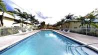 This single-story property showcases a generously sized outdoor pool area that serves as a central amenity and gathering space. The pool itself is expansive and well-maintained, with clear blue water reflecting the surrounding tropical landscape. A wide pool deck provides ample room for relaxation, lined with lounge chairs and seating arrangements positioned to take advantage of the sun and surroundings.

Towering palm trees frame the perimeter of the pool area, creating a lush, tropical atmosphere that enhances the resort-like ambiance. Thatched tiki-style shade structures are strategically placed throughout the deck, offering shaded retreats from the sun. A white perimeter fence encloses the pool area, providing both safety and a sense of privacy.

In the background, a low-rise yellow building is visible, consistent with the single-story nature of the property. The overall environment evokes a relaxed, Florida-style lifestyle, with mature tropical landscaping contributing to a sense of seclusion and warmth. This outdoor space is well-suited for leisure and social activities in a vibrant, sun-filled setting.