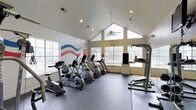 The Stewart Creek fitness center in Frisco is a well-appointed, single-floor workout facility designed to support an active lifestyle. The space is defined by its striking vaulted ceilings that soar to a dramatic peak, creating an open and airy atmosphere that makes the room feel expansive. Large gable windows at the far end of the room flood the interior with abundant natural light, offering pleasant outdoor views while you exercise. A ceiling fan is centrally mounted to promote air circulation throughout the space. The fitness area is equipped with a variety of cardio machines, including stationary bikes, ellipticals, and treadmills, all arranged to take full advantage of the natural light streaming in from multiple windows along the walls. Recessed lighting supplements the natural light, ensuring the space remains bright at all times. A decorative wave mural in patriotic red, white, and blue adds a vibrant artistic touch to one wall, while the "Stewart Creek" name is prominently displayed, reinforcing the community identity. A mounted television and a pull-up/dip station round out the available equipment. The durable fitness flooring throughout ensures both safety and functionality, making this a practical and welcoming space for residents.