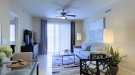 This single-floor residence centers around a bright and welcoming living area designed with a clean, contemporary aesthetic. The room is anchored by a large sliding glass door dressed with vertical blinds and framed by deep blue curtain panels, allowing generous natural light to filter into the space while offering privacy when needed. A ceiling fan with an integrated light fixture is mounted overhead, contributing both functionality and a finishing architectural touch to the room.

The living area is furnished with a light gray sofa accented by decorative throw pillows and paired with a sleek glass-top coffee table resting on a patterned area rug. A large framed abstract artwork adorns one wall, adding visual depth and a sophisticated design element. A floor lamp provides supplemental ambient lighting alongside the natural light from the sliding door.

Adjacent to the living area, a dining space is partially visible, featuring dark-framed chairs that complement the room's modern tone. Tile flooring runs throughout, lending a cohesive, easy-to-maintain surface. The overall ambiance is calm and contemporary, with a neutral color palette punctuated by tasteful decorative accents.