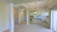This single-floor residence showcases an open-concept design that integrates the living, dining, and kitchen areas into one cohesive, flowing space. Decorative white columns with classical detailing define the transition between zones while maintaining visual openness throughout. A graceful archway frames the entry into the main living area, adding architectural character and depth to the layout.

The kitchen is positioned prominently within the open floor plan and features white cabinetry extending to the upper walls, a granite-topped island that provides additional workspace and casual seating, and white appliances including a refrigerator, range, and built-in microwave. A built-in desk area is neatly integrated into the kitchen perimeter, offering a functional workspace within easy reach.

Crown molding accents the ceilings, complementing the tray ceiling detail visible near the kitchen, which adds dimension overhead. Recessed and pendant lighting illuminate the space, with a statement hanging fixture positioned above the island. Neutral-toned walls and light carpeting in the living area create a warm, understated ambiance. A dedicated laundry area is visible through an adjacent doorway, adding practical utility to the home's well-organized layout.