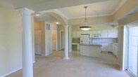 This single-floor residence showcases an open-concept design that integrates the living, dining, and kitchen areas into one cohesive, flowing space. Decorative white columns with classical detailing define the transition between zones while maintaining visual openness throughout. A graceful archway frames the entry into the main living area, adding architectural character and depth to the layout.

The kitchen is positioned prominently within the open floor plan and features white cabinetry extending to the upper walls, a granite-topped island that provides additional workspace and casual seating, and white appliances including a refrigerator, range, and built-in microwave. A built-in desk area is neatly integrated into the kitchen perimeter, offering a functional workspace within easy reach.

Crown molding accents the ceilings, complementing the tray ceiling detail visible near the kitchen, which adds dimension overhead. Recessed and pendant lighting illuminate the space, with a statement hanging fixture positioned above the island. Neutral-toned walls and light carpeting in the living area create a warm, understated ambiance. A dedicated laundry area is visible through an adjacent doorway, adding practical utility to the home's well-organized layout.