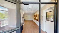 Located at 200 Garden St in South Burlington, Vermont, this single-floor hallway and common corridor space offers a clean, modern aesthetic designed to welcome and orient visitors and residents alike. The interior features warm wood-tone luxury vinyl plank flooring throughout, complemented by crisp white walls and a drop-panel ceiling with recessed lighting that provides bright, even illumination across the space.

A central elevator with stainless steel doors serves as a functional focal point in the main hallway, flanked by wall-mounted sconces and framed by thoughtfully selected artwork. The corridors are adorned with an impressive rotating display of curated artwork, including large-scale abstract paintings in vibrant hues and striking geometric wood-panel compositions, lending the space a gallery-like quality. Tufted leather benches are positioned throughout, offering comfortable waiting areas for residents and guests.

The building's exterior, visible from the accessible ramp and entry points, showcases a contemporary facade with horizontal siding and large windows. The entrance features a covered walkway with black metal railings overlooking a well-maintained parking area surrounded by lush Vermont greenery. The overall ambiance is polished and welcoming, balancing functional circulation with an elevated aesthetic that sets a refined tone for the building.