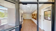 Located at 200 Garden St in South Burlington, Vermont, this single-floor hallway and common corridor space offers a clean, modern aesthetic designed to welcome and orient visitors and residents alike. The interior features warm wood-tone luxury vinyl plank flooring throughout, complemented by crisp white walls and a drop-panel ceiling with recessed lighting that provides bright, even illumination across the space.

A central elevator with stainless steel doors serves as a functional focal point in the main hallway, flanked by wall-mounted sconces and framed by thoughtfully selected artwork. The corridors are adorned with an impressive rotating display of curated artwork, including large-scale abstract paintings in vibrant hues and striking geometric wood-panel compositions, lending the space a gallery-like quality. Tufted leather benches are positioned throughout, offering comfortable waiting areas for residents and guests.

The building's exterior, visible from the accessible ramp and entry points, showcases a contemporary facade with horizontal siding and large windows. The entrance features a covered walkway with black metal railings overlooking a well-maintained parking area surrounded by lush Vermont greenery. The overall ambiance is polished and welcoming, balancing functional circulation with an elevated aesthetic that sets a refined tone for the building.