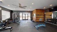 The fitness center at 1101 Pure St in Saint Peters, Missouri, is a well-appointed, single-floor facility designed to support a comprehensive workout experience. The space is divided into two interconnected workout areas, each with a distinct character and equipment selection.

The main workout area features dark gray walls, large-format tile flooring, and a standout horizontal wood-plank accent wall branded with the "Pure" logo, adding a warm, modern aesthetic to the otherwise industrial-style space. A large ceiling fan with recessed lighting ensures proper ventilation and illumination. Glass double doors lead to an outdoor area, filling the room with natural light. Equipment here includes a cable machine, rowing machine, adjustable benches, and a dumbbell rack.

The secondary workout area features softer sage green walls contrasted by a dark charcoal accent wall, wall-to-wall mirrors, and additional recessed lighting supported by a ceiling fan. This section houses a comprehensive dumbbell rack with kettlebells, adjustable benches, cardio machines including treadmills, and a multi-function cable/pulley system. Windows with blinds allow controlled natural light throughout. A water fountain and wall-mounted television add convenience and comfort to the overall facility.