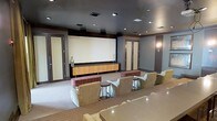 This single-floor screening room is designed to deliver a premium home theater experience. At the front of the room, a large projection screen serves as the focal point, framed by a sleek, dark-painted ceiling that enhances contrast and minimizes light reflection during screenings. Recessed lighting is strategically placed throughout the ceiling to provide ambient illumination when the room is not in use for viewing, while wall sconces add a warm accent glow.

A long, warm-toned wood media credenza sits beneath the screen, providing storage and housing for media equipment. Surround sound speakers are mounted along the upper perimeter of the walls, ensuring full audio immersion. The seating arrangement features multiple plush lounge chairs and upholstered benches positioned in a tiered layout, offering unobstructed sightlines to the screen from every seat.

The walls are finished in a muted gray tone, complementing the dark ceiling and neutral carpeted flooring. Framed double doors with warm wood trim flank both sides of the room, adding a refined, architectural detail. Small side tables are placed between seating areas for added convenience. The overall ambiance is sophisticated, functional, and purpose-built for entertainment.