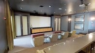 This single-floor screening room is designed to deliver a premium home theater experience. At the front of the room, a large projection screen serves as the focal point, framed by a sleek, dark-painted ceiling that enhances contrast and minimizes light reflection during screenings. Recessed lighting is strategically placed throughout the ceiling to provide ambient illumination when the room is not in use for viewing, while wall sconces add a warm accent glow.

A long, warm-toned wood media credenza sits beneath the screen, providing storage and housing for media equipment. Surround sound speakers are mounted along the upper perimeter of the walls, ensuring full audio immersion. The seating arrangement features multiple plush lounge chairs and upholstered benches positioned in a tiered layout, offering unobstructed sightlines to the screen from every seat.

The walls are finished in a muted gray tone, complementing the dark ceiling and neutral carpeted flooring. Framed double doors with warm wood trim flank both sides of the room, adding a refined, architectural detail. Small side tables are placed between seating areas for added convenience. The overall ambiance is sophisticated, functional, and purpose-built for entertainment.