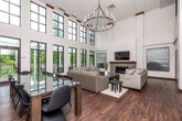 Located at 3500 Edgewood Rd NE in Cedar Rapids, Iowa, this single-floor space makes a dramatic architectural statement with its soaring two-story ceilings and an expansive wall of floor-to-ceiling gridded windows that flood the interior with abundant natural light. The open-concept layout seamlessly connects the living, dining, and kitchen areas, creating a cohesive and functional flow throughout the space.

The living area is anchored by a sleek stone-surround linear fireplace with a wood mantel and a wall-mounted flat-screen television above. Neutral-toned furnishings, a glass-top coffee table, and a soft area rug contribute to the refined, contemporary aesthetic. A statement chandelier with a wrought-iron frame and candle-style lights hangs above the large dining table, which comfortably seats multiple guests. Rich, dark hardwood flooring runs throughout, adding warmth and contrast to the bright white walls.

The kitchen features warm wood cabinetry, stainless steel appliances including a refrigerator, range, and microwave, and a generous white-topped peninsula with bar seating — ideal for casual dining or entertaining. Light tile flooring in the kitchen area transitions smoothly to the hardwood beyond. French doors open to an outdoor deck, offering views of the surrounding lush greenery and extending the living experience outdoors.