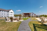 Located at 1905 Riverside Pkwy in Douglasville, Georgia, this residential community is defined by its thoughtfully designed outdoor amenity grounds. The exterior spaces serve as the centerpiece of the property, offering a generous central lawn area lined with Adirondack chairs positioned to encourage relaxation and social gathering. Winding gravel pathways connect the various outdoor zones, creating a cohesive and walkable environment throughout the community.

A standout feature is the bold, wood-slatted pergola structure that provides shaded outdoor space alongside a grilling station, making it ideal for outdoor dining and entertainment. Adjacent to the pergola, a stone-ringed fire pit surrounded by Adirondack chairs creates an inviting gathering spot for residents. A dedicated lawn game area with cornhole boards adds a recreational element to the grounds.

The property is framed by multiple multi-story residential buildings finished in white and light gray exteriors with pitched rooflines, contributing to a clean, modern aesthetic. The landscaping throughout features manicured turf, mulched garden beds, ornamental plantings, and mature trees along the perimeter, providing a natural backdrop. Ample surface parking is accessible via a well-maintained driveway, and the surrounding wooded treeline offers a sense of privacy and natural scenery under the open Georgia sky.