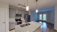 Located at 401 E Stone Ave in Greenville, SC, this single-floor residence offers a thoughtfully designed open-concept layout with contemporary finishes throughout. The kitchen serves as the focal point of the main living area, featuring a large granite-topped island with a double undermount sink and pendant lighting overhead. Gray shaker-style cabinetry lines the walls alongside a white subway tile backsplash, while stainless steel appliances—including a refrigerator, electric range, and built-in microwave—complete the modern aesthetic. Track lighting provides additional illumination throughout the kitchen. Light wood-style flooring runs through the main living and kitchen areas, creating a warm, cohesive feel that extends toward French doors offering views of greenery outside. The bedroom areas feature plush carpeting and neutral gray walls, with ample natural light from large windows with blinds. Closet access is conveniently integrated into the bedroom spaces. The bathroom is finished with a granite-topped vanity, large mirror, subway tile tub surround with a curved shower rod, and tile flooring, maintaining the consistent modern aesthetic found throughout the unit.