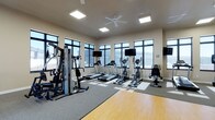 This fitness center is designed to support a comprehensive workout experience within a single, open-plan space. The room is characterized by large, dark-framed windows lining the walls, allowing abundant natural light to fill the interior while offering views of the surrounding exterior. A ceiling fan centered overhead complements the recessed panel lighting, contributing to a comfortable and well-ventilated atmosphere.

The flooring is a combination of durable hardwood-style material and fitted carpet, delineating areas suited for different types of exercise. Cardio equipment is prominently featured, including multiple treadmills and elliptical machines, each positioned facing mounted flat-screen televisions for an engaging workout experience. A multi-station strength training machine is situated along one side of the room, offering a variety of resistance-based exercises. Additional cardio options, such as a stationary bike, round out the equipment selection.

The neutral, light-toned walls and high ceiling panels contribute to an open and airy ambiance. The layout is thoughtfully organized, allowing for comfortable movement between stations while maximizing the usable floor space throughout the room.
