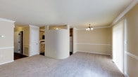 This two-floor space showcases a thoughtfully designed open-concept main level that seamlessly connects the living, dining, and kitchen areas. A standout architectural feature is the curved, cylindrical dividing wall that serves as a subtle partition between the living space and the kitchen, adding a unique visual element to the layout. The living and dining areas are finished with neutral-toned wall-to-wall carpeting, contributing to a warm and comfortable atmosphere. Crown molding lines the upper edges of the walls, lending a refined touch to the interior. An elegant chandelier with multiple light fixtures anchors the dining area, providing both functional and ambient lighting. The kitchen features light wood cabinetry, a white refrigerator, and dark flooring, offering a practical workspace that remains visually connected to the main living area. Warm beige tones throughout the walls create a cohesive and inviting palette across the space. A white interior door near the kitchen provides access to an additional room or utility space. The overall design balances open, airy living with gentle structural definition, making effective use of the available floor space across two levels.