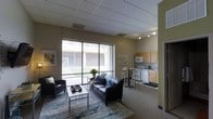 This single-floor space offers an open-concept layout that seamlessly connects the living area and kitchen, creating a cohesive and functional environment. The living area is anchored by a dark leather sofa and armchair arrangement, complemented by a patterned area rug and a glass-top coffee table. A large floor-to-ceiling window spans the width of the living space, allowing ample natural light to filter in while offering a view of an adjacent outdoor courtyard area. The drop-panel ceiling features recessed and track lighting, providing versatile illumination throughout. The kitchen, situated toward the rear of the space, is outfitted with light wood cabinetry, a white electric range, an over-the-range microwave, and a compact sink with counter workspace. A small bistro-style table with bar stools is positioned at the kitchen's edge, serving as a casual dining area. The flooring transitions from carpet in the living zone to tile in the kitchen. Neutral taupe walls and understated décor contribute to a clean, practical, and welcoming ambiance throughout the unit.